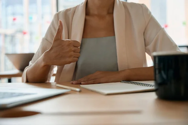 Woman in office shows hand gesture that everything is fine. She is sitting at table with documents, cup and laptop