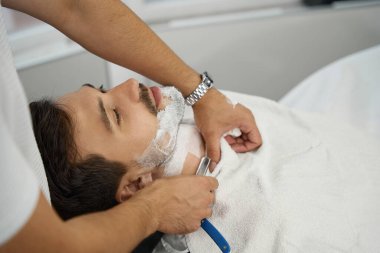 Sakin esmer adam traş olurken berber koltuğuna uzanıyor.