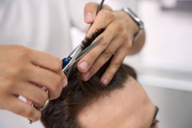 Berberin makas ve tarak kullanarak müşterisinin koyu renk saçlarını keserken çekilmiş fotoğrafı.