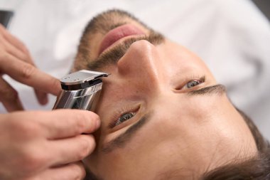 Esmer adamın yakın plan fotoğrafı berber dükkanında sakalını budarken ve elektrikli makasla şekillendirirken çekilmiş.