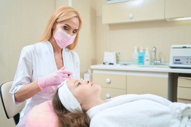 Doktor kozmetik uzmanı kozmetik muayenehanesinde sandalyede yatan genç bir kızın alnına iğne yapıyor.