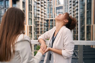 Ofis balkonunun korkuluklarına yaslanan ve iş arkadaşının elini tutan neşeli bir şirket çalışanı.