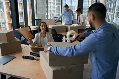 Şirket çalışanı elinde belgelerle dizüstü bilgisayarında oturmuş, karton kutuyu koli bandıyla mühürleyen iş arkadaşına bakıyor.