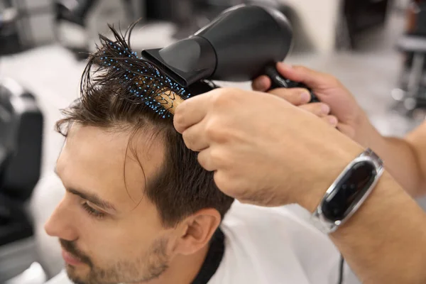 Cropped close-up shot of dark-haired gentleman having his hair styled and dried at barbershop