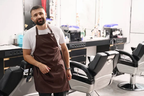 Uśmiechnięty Fryzjer Fartuchu Opierając Się Fotelu Podczas Pozowania Salonie Fryzjerskim — Zdjęcie stockowe