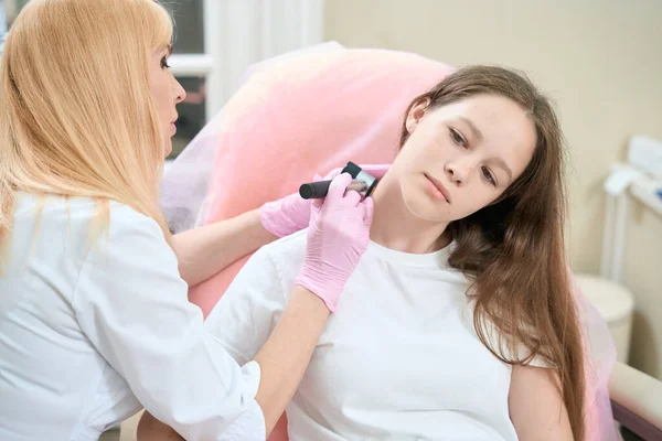戴着粉色手套的漂亮女医生肿瘤学家在诊所用皮肤科镜检查少女的颈部 — 图库照片