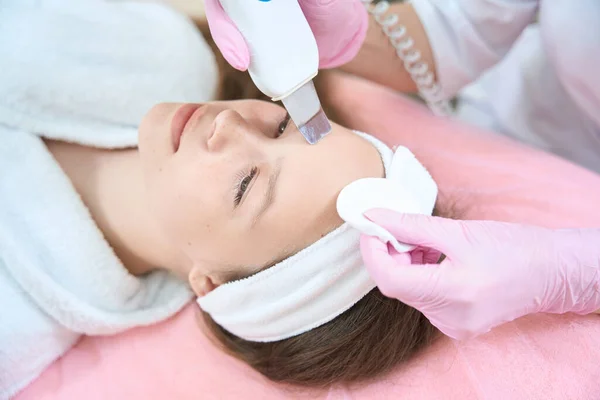 Teenage girl lies in a pink armchair during an ultrasonic cleaning with a scrubber in cosmetology clinic. Face close-up