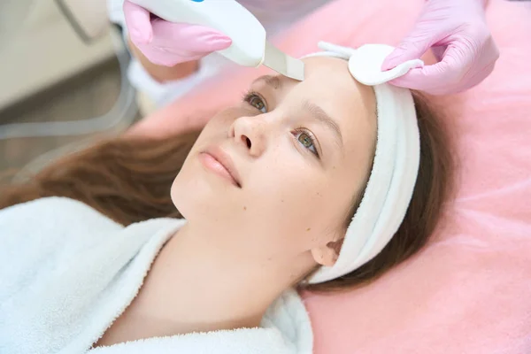 ピンクの手袋の女性皮膚科医は美容クリニックで女の子の顔にスクラバーで超音波洗浄を行います — ストック写真