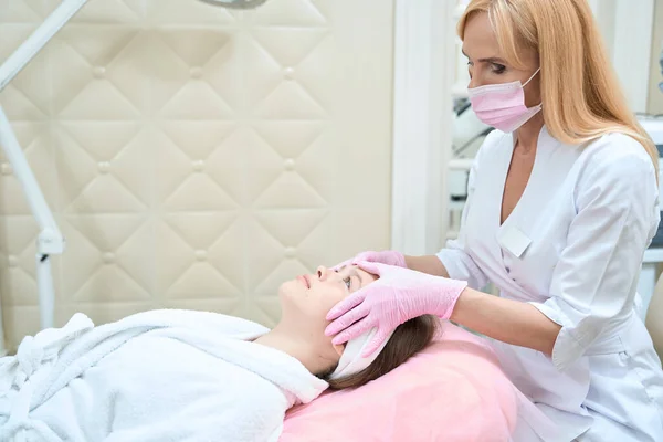 Focused female beautician in protective mask holds face of girl who lies in pink chair. Cosmetological procedures in beauty salon