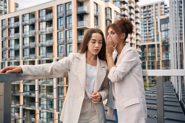 Company employee whispering something in ear of her female coworker on outdoor balcony