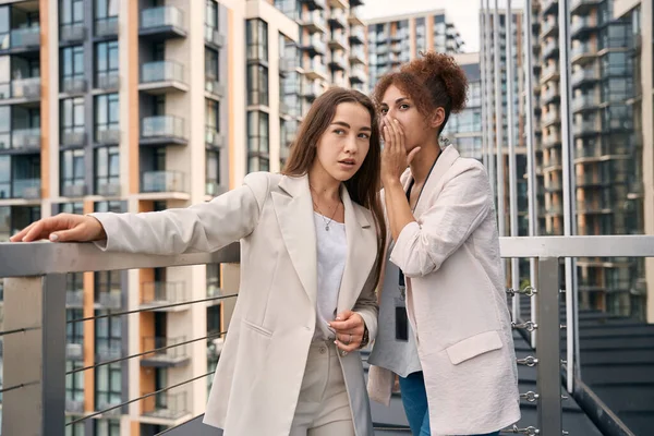 Company worker whispering something in ear of her female coworker on balcony