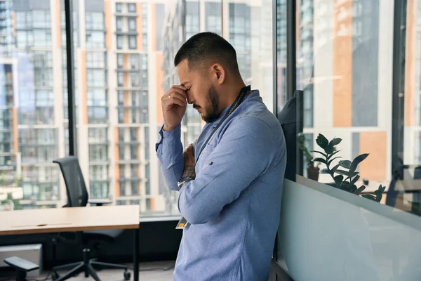 Side view of corporate employee leaning against glass office wall while pinching bridge of his nose with hand