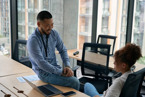 Joyful company manager sitting on office desk and talking to his coworker