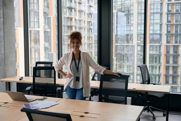 Smiling confident female entrepreneur standing at her office desk and leaning on mesh chair backrest