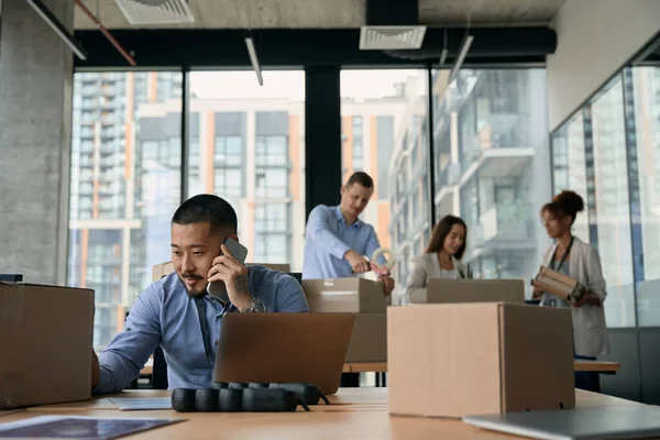 彼のチームは段ボール箱を梱包しながら 若い企業のマネージャーは 電話会話中にラップトップに座っていた — ストック写真