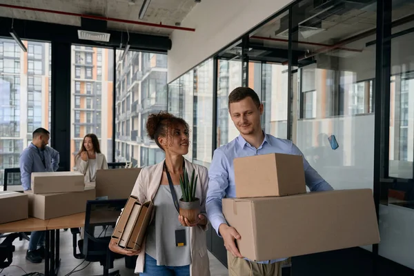 Corporate employee holding file folders and potted plant in hands and looking at colleague with cardboard boxes