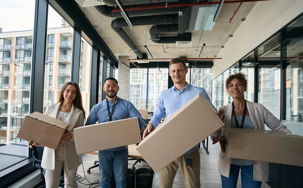 Cheerful corporate workers standing in office and holding sealed cardboard boxes in hands