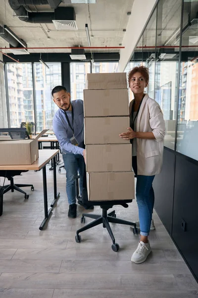 Full-length portrait of corporate workers pulling swivel chair with pile of sealed cardboard boxes