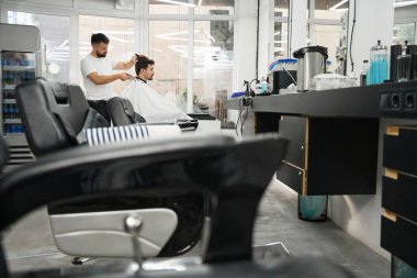 Berber dükkanındaki müşterinin koltuktaki yan görüntüsü ve arkasındaki profesyonel kuaför saç kesimiyle meşgul.