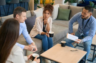 Multiethnic team of serious entrepreneurs communicating at coffee table during business meeting clipart