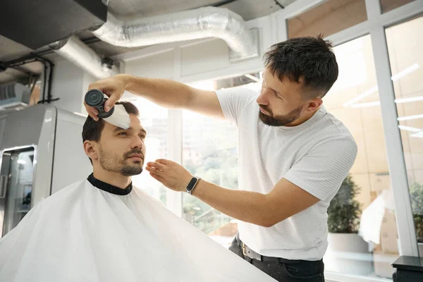 Focused barber looking at client while carefully cleaning his face from the hair remainders with a brush