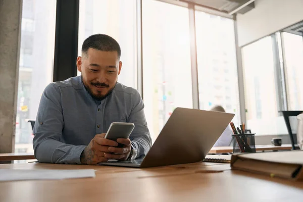 Uśmiechnięty Pracownik Korporacyjny Siedzący Laptopie Patrzący Smartfona Jego Rękach — Zdjęcie stockowe