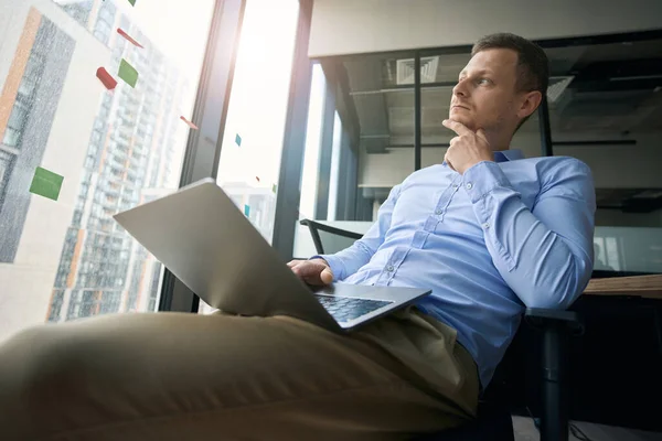 Serious thoughtful young office manager with laptop sitting in chair by window