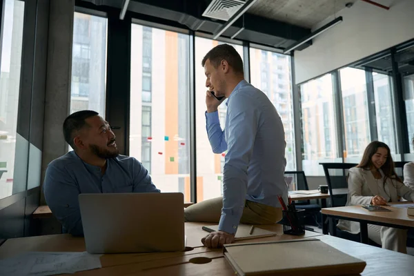 Joyful corporate employee seated at portable computer watching his colleague talking on smartphone