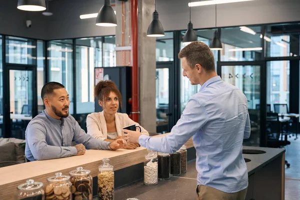 Friendly young company worker treating his colleagues to coffee in office kitchen