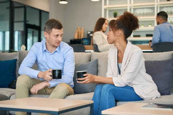 Serious young man talking to woman seated on sofa in co-working space