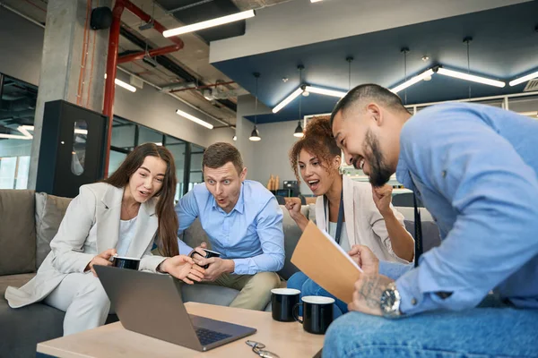 Multiracial group of surprised business colleagues sitting around coffee table and watching video on laptop