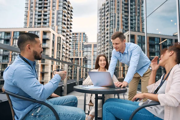 Young corporate worker with his laptop communicating with his coworkers seated on table outside