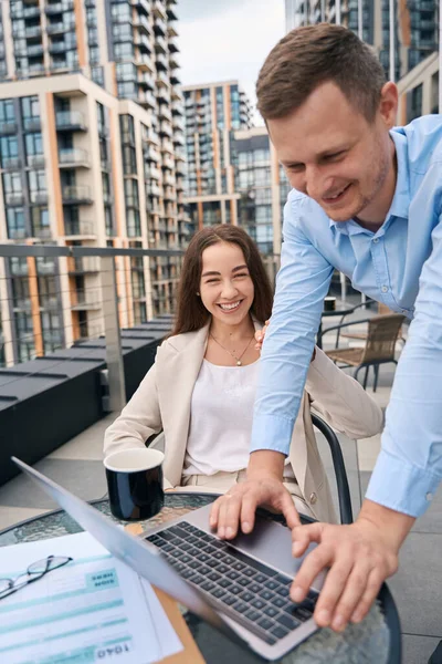Cheerful young corporate employee typing on laptop supported by his female colleague