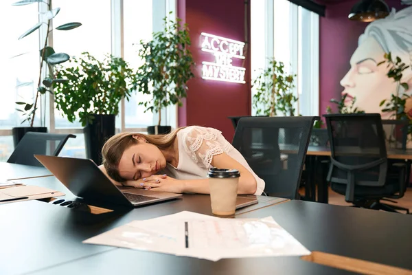 Weary young corporate employee napping on desk before laptop during coffee break