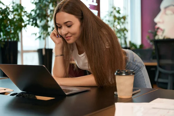 Smiling young company employee leaning on desk while looking at laptop screen