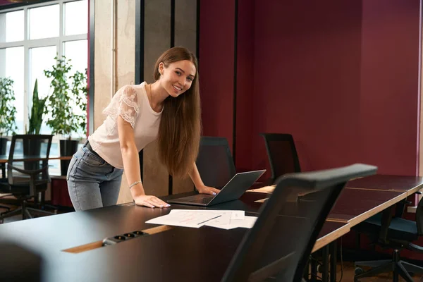 Smiling pleased young company employee leaning on office desk before portable computer