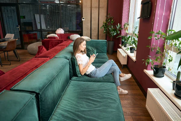 彼女の携帯電話を使用してコワーキングスペースでソファに座って焦点を当てた女性の側面図 — ストック写真