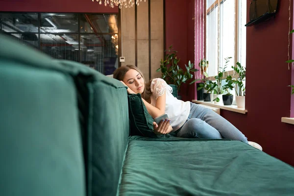 リラックスした若い女性は快適なソファに横になり 携帯電話でテキストメッセージを読む — ストック写真