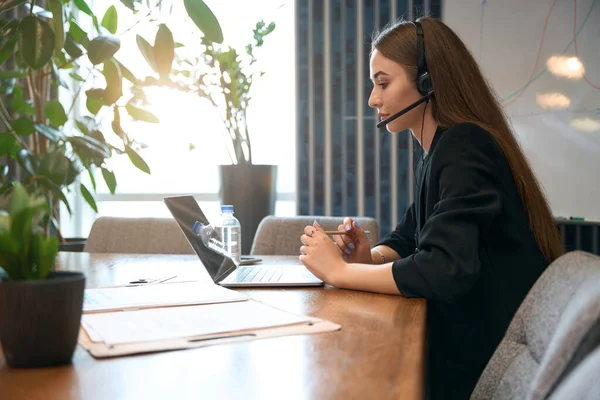 Side view of Internet shop worker in headset seated at desk staring at laptop screen