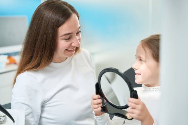 Memnun bir pedodontiste bakarken elinde ayna tutan neşeli genç bir hasta.