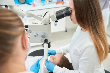 Ciddi odaklı dişçi, kadın hemşirenin yardımıyla mikroskop altında çocuğun ağız boşluğunu inceliyor.
