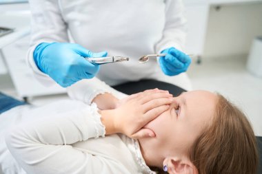 Küçük kız dişçi aynası ve cımbızı olan bir doktorun huzurunda elleriyle ağzını kapatıyor.