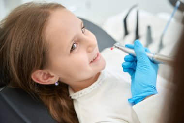 Neşeli genç hasta dişçi koltuğunda yatıp pediatrik dişçiye bakıyor.