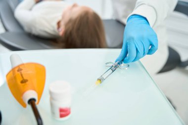 Nitrile eldivenli pediatrik dişçinin kesilmiş fotoğrafı hastanın yanına oturmuş diş şırıngasına uzanıyor.