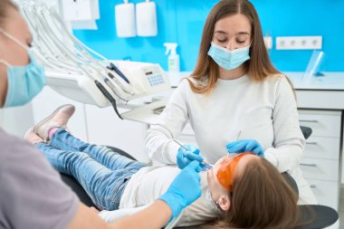 Azot eldivenli stomatolog, yanak retraktörü ve diş araştırması kullanarak küçük hasta dişlerini inceliyor.