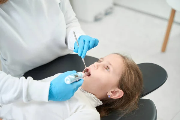 Pediatric dentist in nitrile gloves injecting anesthetic into gum of little girl using dental syringe gun