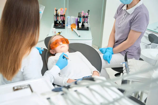 Little patient in safety glasses lying in dental chair surrounded by pediatric dentist and nurse