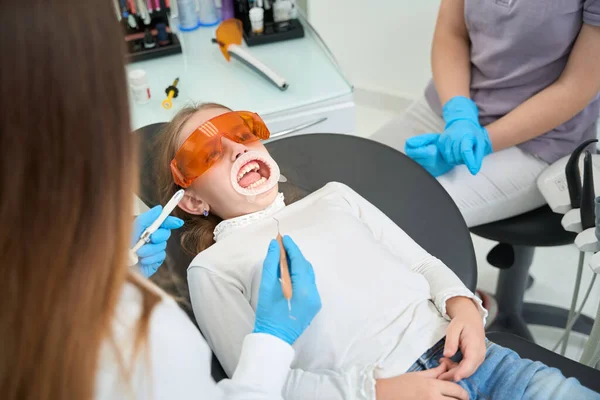面颊牵引器的小病人躺在被牙科医生和护士包围的椅子上 — 图库照片