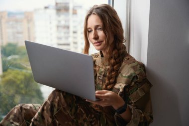 Düşünceli asker hanım, dizleri dizlerine eğilmiş bir dizüstü bilgisayar kullanarak pencerenin kenarına oturur.