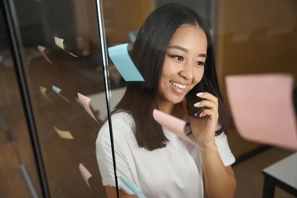 笑顔のアジアの女性のクローズアップ肖像画は 現代のオフィスのガラスのドアの近くに立っている間 スマートフォンで会話をしています — ストック写真